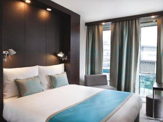 Modern hotel bedroom with a large bed accented by teal and white bedding, flanked by sleek bedside lighting, set against a backdrop of a city view through large windows